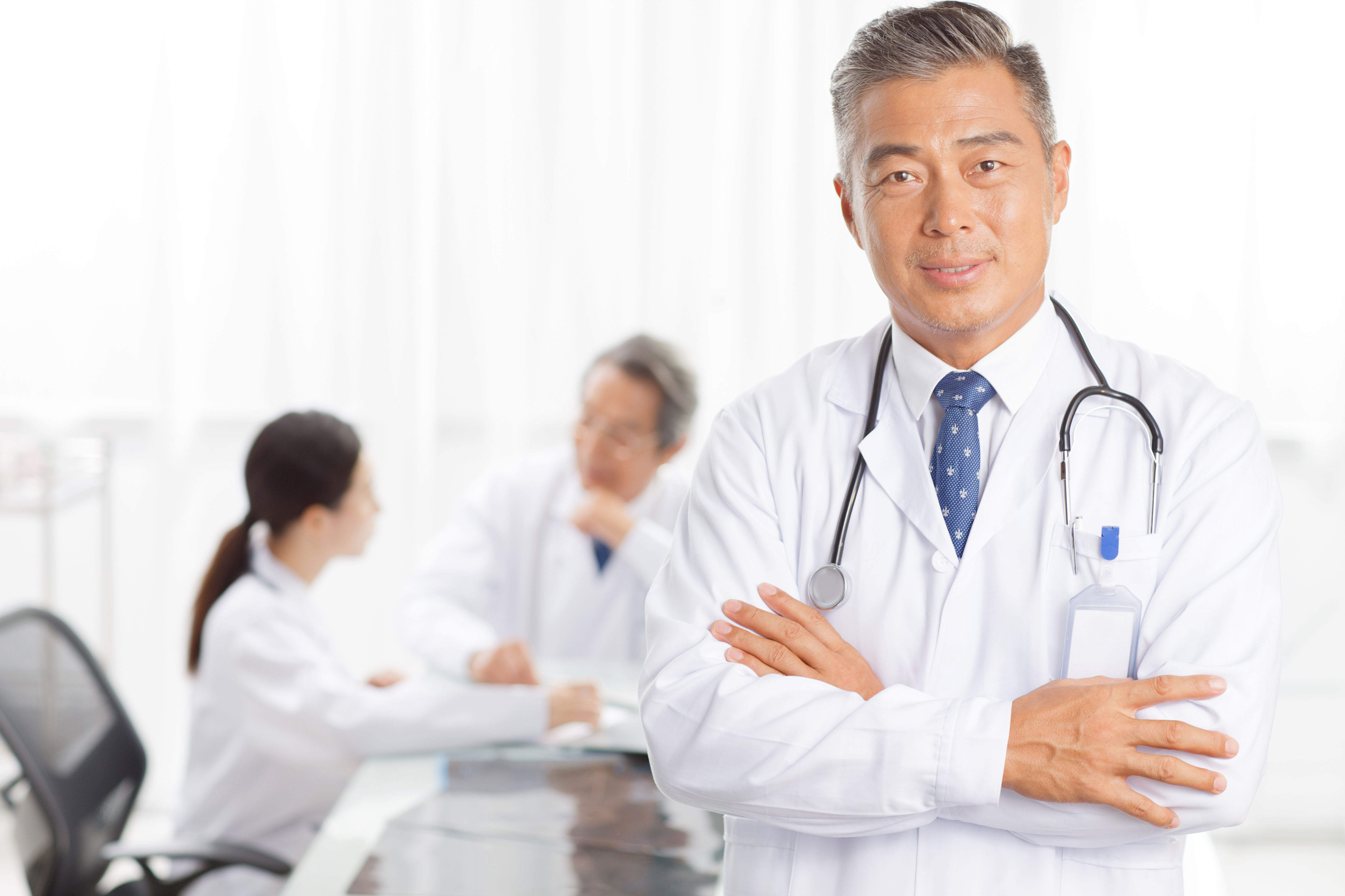 A doctor wearing a white coat stands at the foreground with their arms crossed while two other doctors sit in conversation in the background.